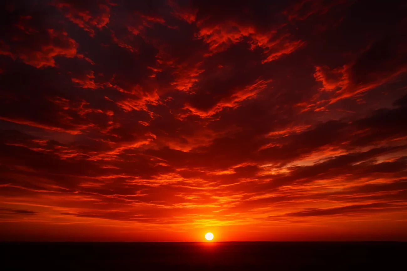 Atardecer sobre un paisaje abierto con el sol tocando el horizonte y nubes teñidas de rojo, naranja y púrpura sobre el cielo que cambia de color.