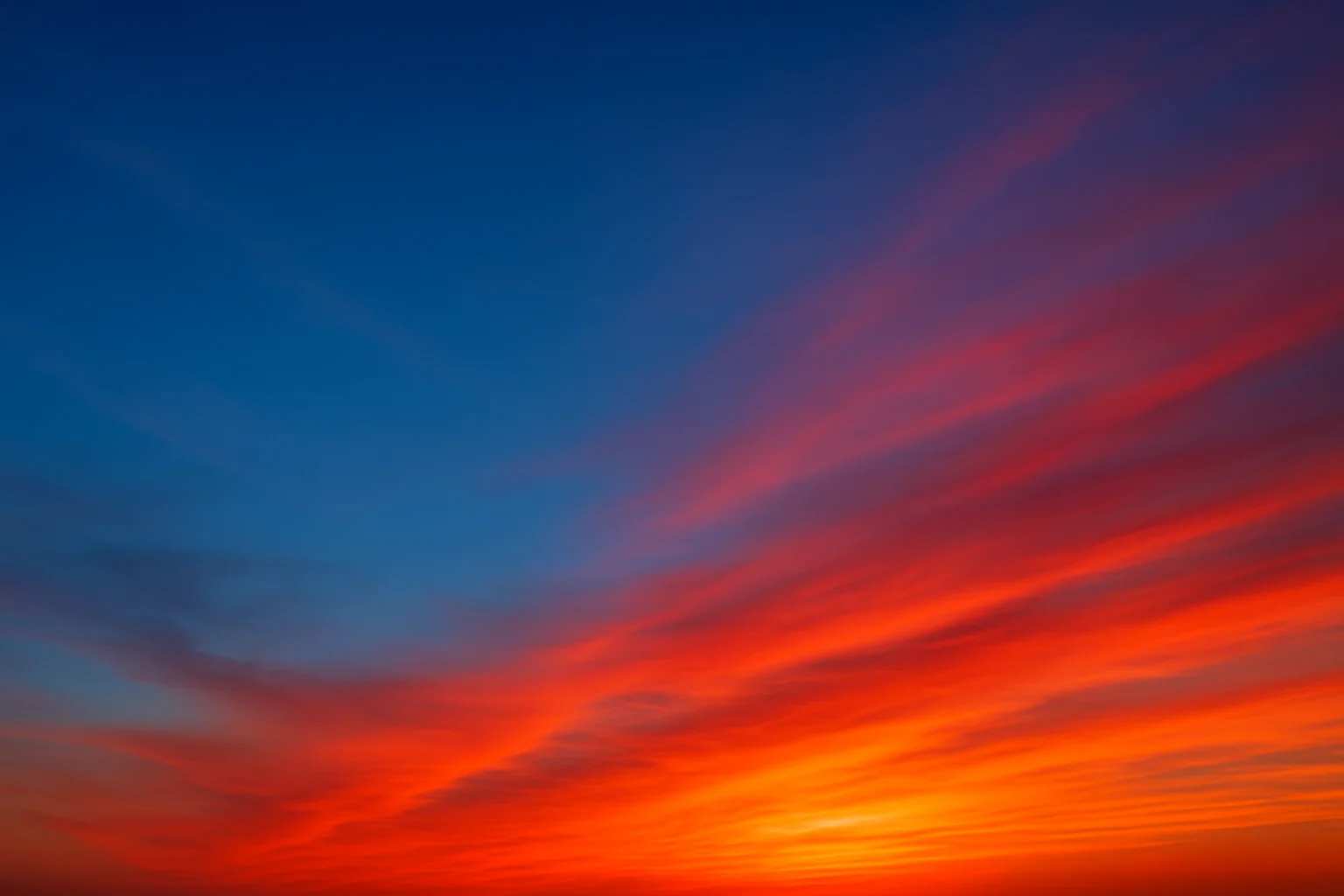 Cielo al atardecer con un degradado de azul a rojo, mostrando nubes que se tiñen de colores cálidos mientras el sol se oculta en el horizonte.