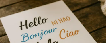 Hoja de papel con la palabra “hola” escrita en distintos idiomas, sobre una mesa de madera rústica. Al fondo se ve una planta en maceta y una manta de lana.